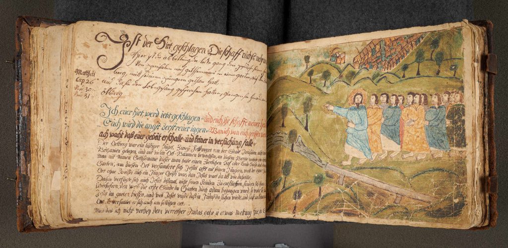 Aerial view of the Ludwig Dening Manuscript, opened and laid flat on a table. Left page includes text in German that has been handwritten in brown, blue, and red ink on the page. Pages are centuries old and show natural yellowing, but the text remains legible. Right page depicts a drawing of the passage on the left, which features Jesus and his followers, walking ahead and through some hills. Image is in full color, including green hills, trees, and a blue river. Each person is in varied colored robes including blue, yellow, and red.