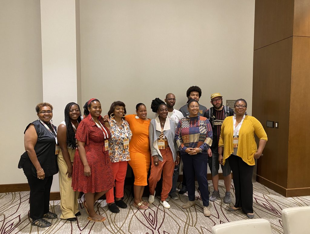 Group photo of 11 participants of this summer's Omeka S workshop at Jackson State. Every person is smiling. In the front row from left to right, there is a woman in a black top and black pants, a woman in a white top and tan pants, a woman in a red dress, a woman in a floral patterned top and red pants, a woman in an orange dress, a woman with a grey sweater and burnt orange pants, a woman with a geometric print top and blue pants, and a woman with a yellow sweater and black pants. In the back row from left to right is a man with a white top, man with a grey top, and man with a striped top and shorts. 