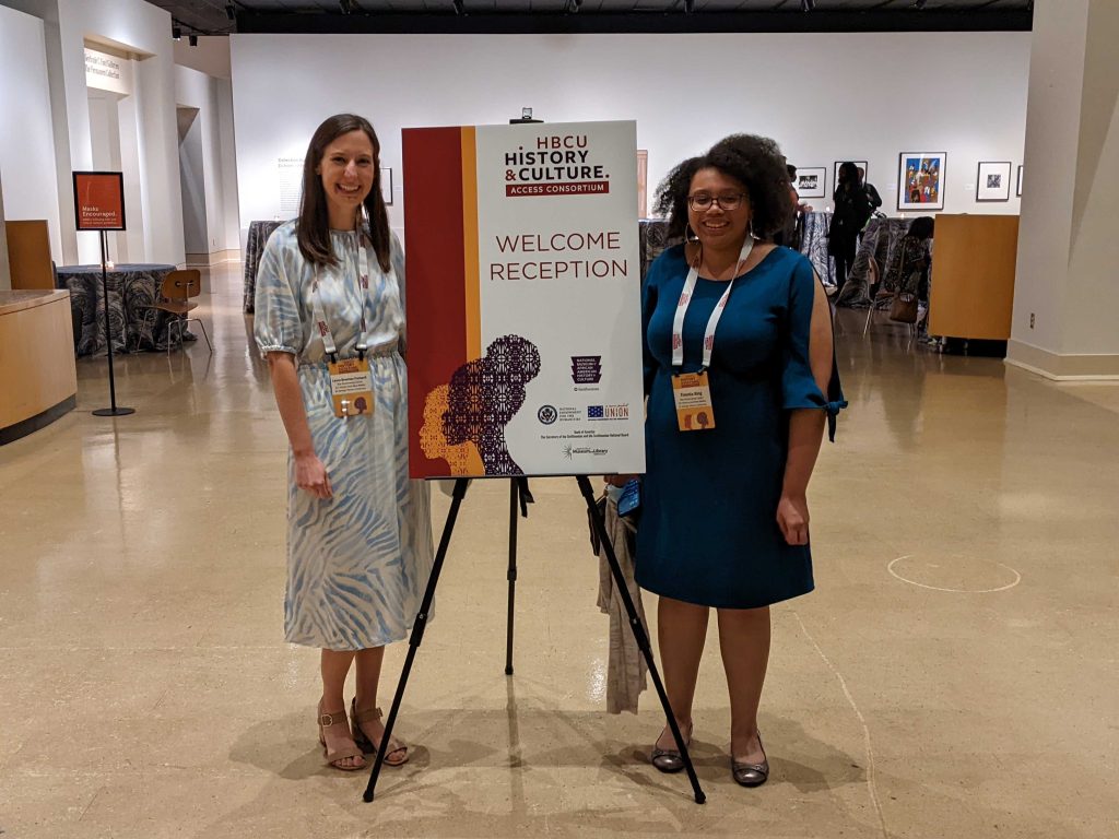 Photo taken at the Welcome Reception for the HBCU History and Culture Access Consortium (HCAC) this summer at Jackson State. In the middle of the photo is the welcome poster that says what the event is (HCAC Welcome Reception). Poster features one gold and one purple silhouette in the foreground of the poster over a red and gold vertical stripe. Flanking the poster's left is Laura Brennan Fretwell, in a white and blue dress, and Timmia King flanks the poster's right in a blue dress.  