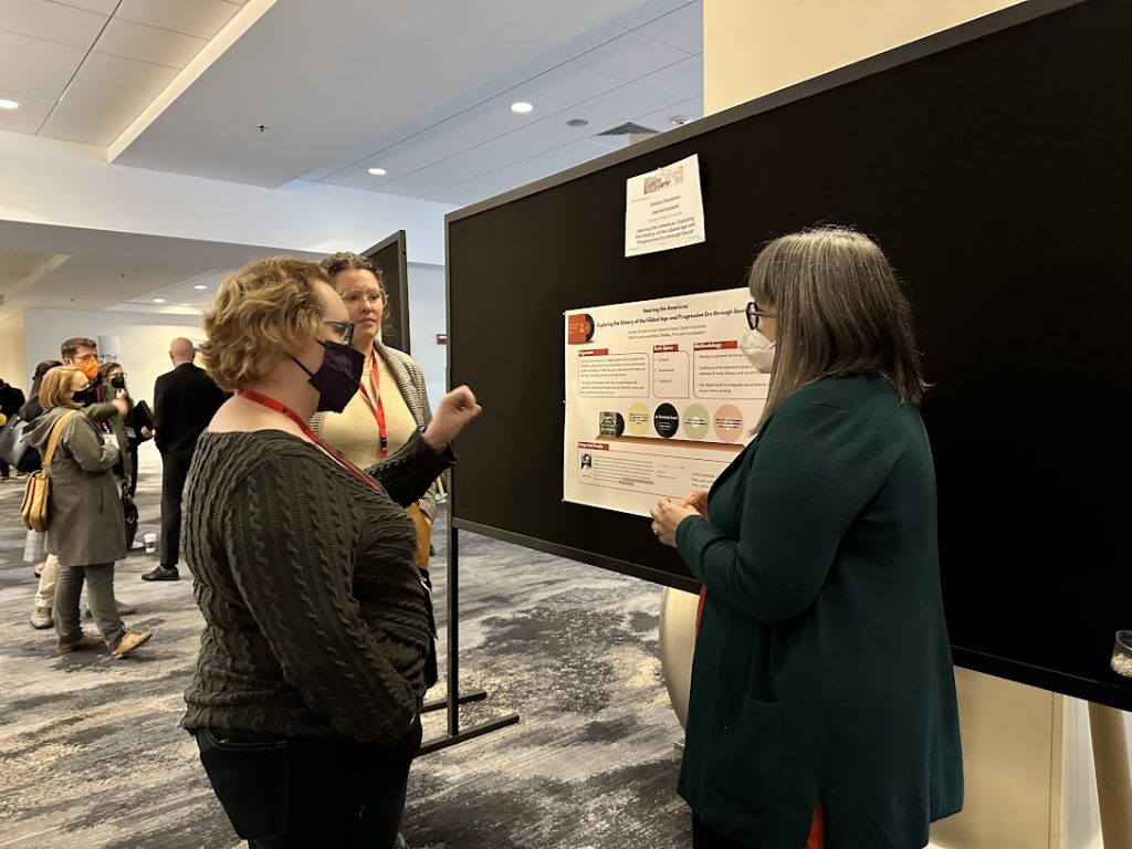 Photo taken at the AHA poster session of Jessica and Jayme flanking their poster about Hearing the Americas, engaged in conversation with a historian interested in learning about the project.