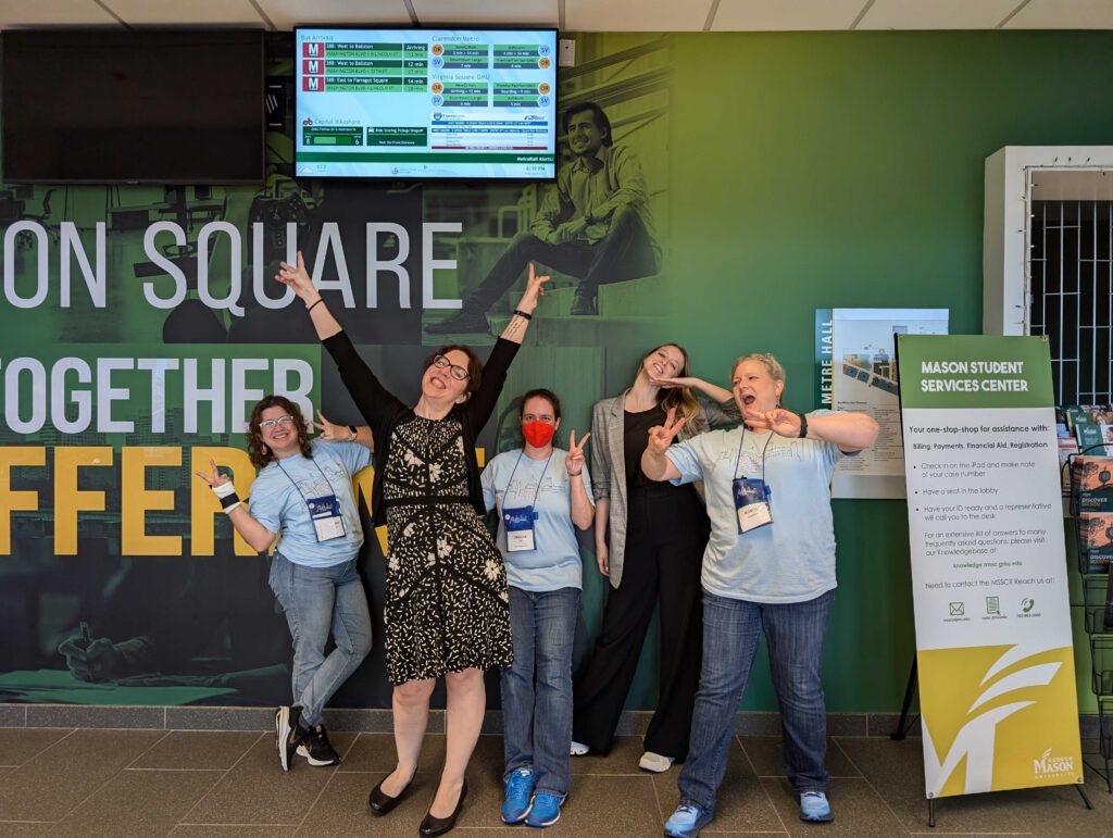 5 Women standing in front of a green wall, 4  with their hands signaling "V" for "Victory" and the 5th with her hand under her chin.